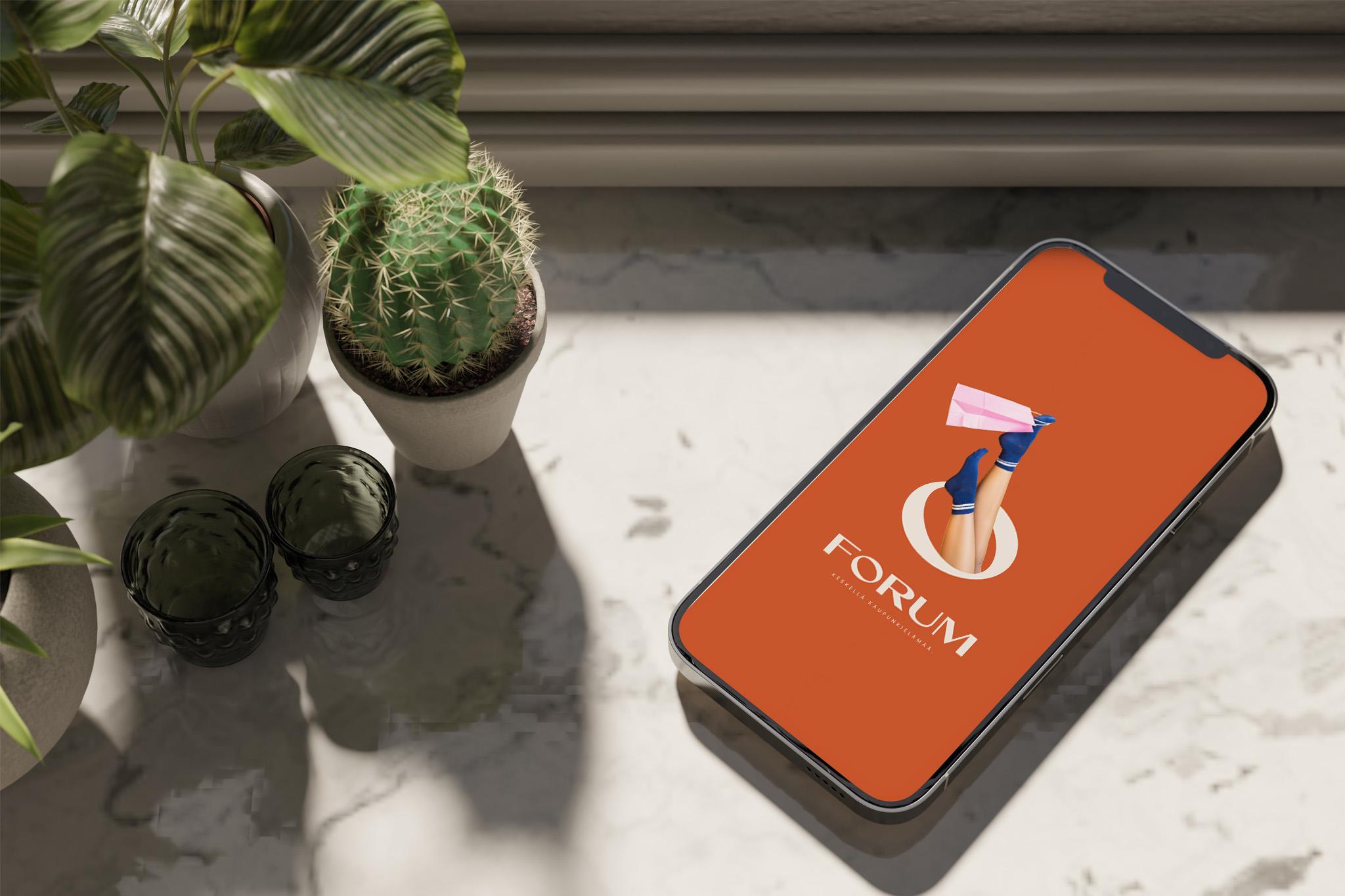 Älypuhelin näyttää Kauppakeskus Forumin mobiilisovelluksen tai mobiilikampanjan oranssilla taustalla, jossa on Forumin logo ja kuva jaloista ostoskassin kanssa.